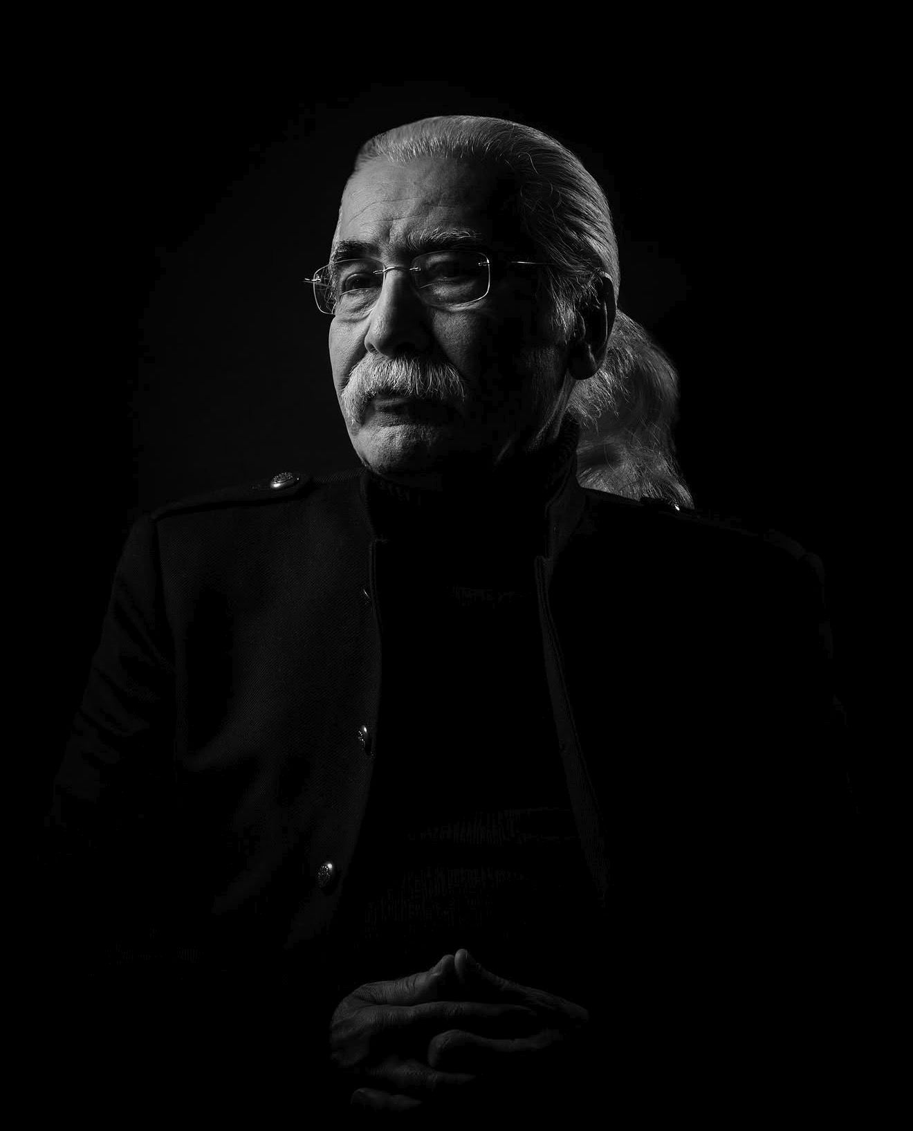 Iraj Jannatie Ataie seated in near darkness, one side of his face lit, hands clasped, wearing rimless glasses.